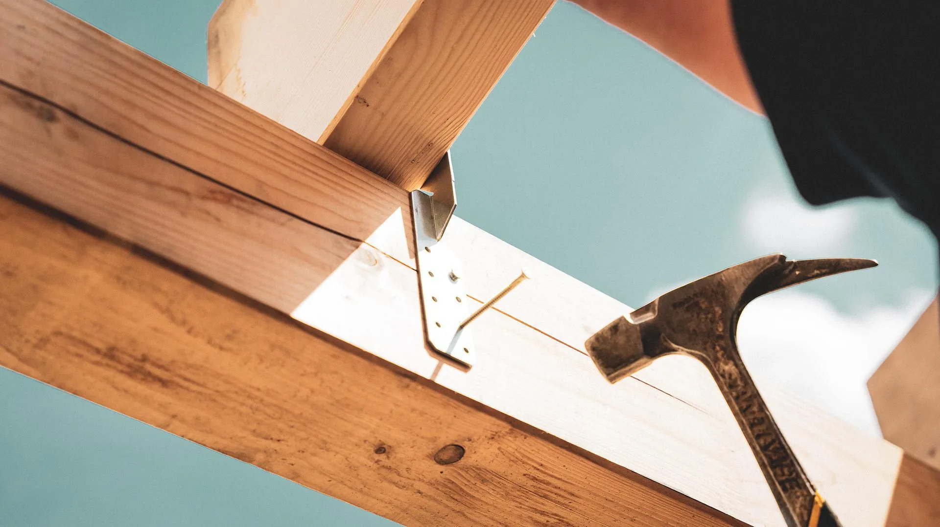 Photo of a builder nailing a bracket into place, a blue sky overhead.